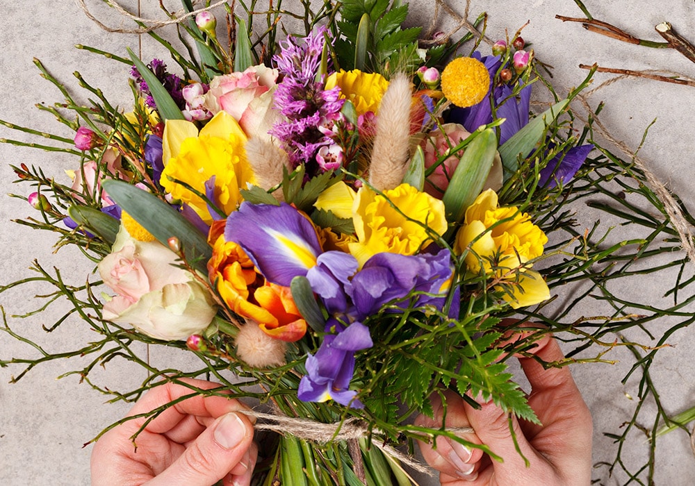 Colorful bouquet with yellow, purple, and pink flowers, held by two hands, against a light gray background, surrounded by scattered green stems and twine.